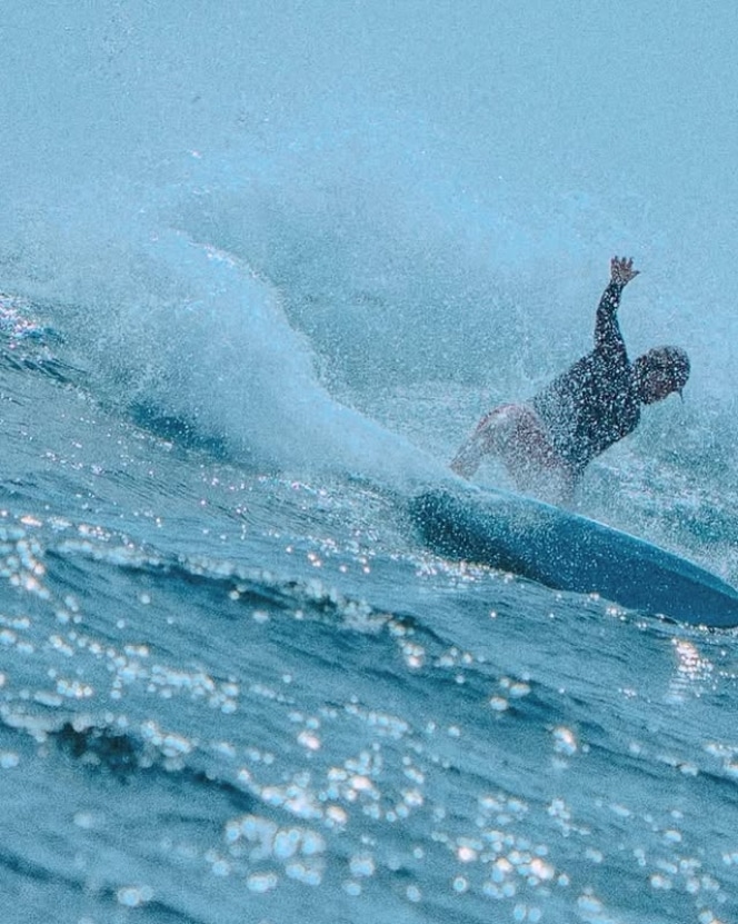 A surfer wearing a black wetsuit loses his balance and falls off his blue surfboard as a wave crashes behind him, sending water flying into the air next to other surfboards riding the swell.
