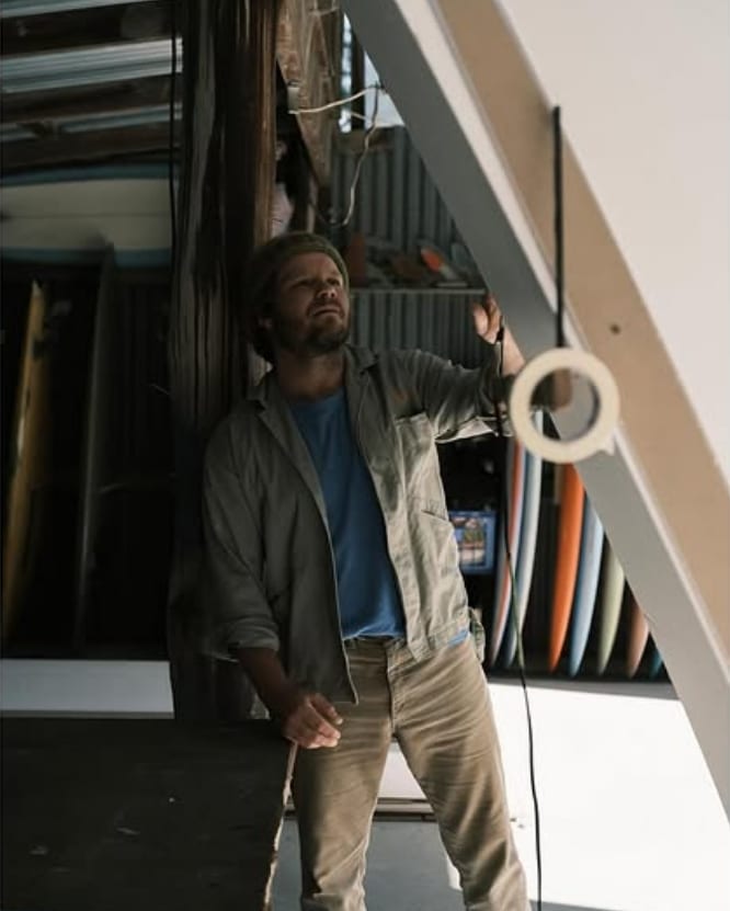 A casually dressed man stands inside, reaching out to touch a white wall or structure. Several vibrant surfboards are stacked vertically in the background, evoking the atmosphere of a surf store or workshop.